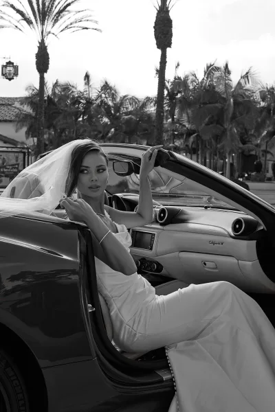 Bridal portrait of a bride in a convertible wearing a strapless satin wedding dress and veil, with bracelet and necklace, palm trees beyond the street
