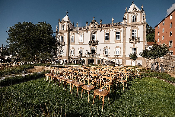 Ceremony setup with outdoor wedding ceremony setup, rows of wood cross back chairs and programs on a lawn before a historic estate and stone stairs.