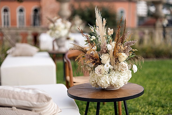 Wedding centerpiece with white rose blooms, hydrangea, and pampas grass in a low bowl on a round wood table beside a lounge sofa outdoors
