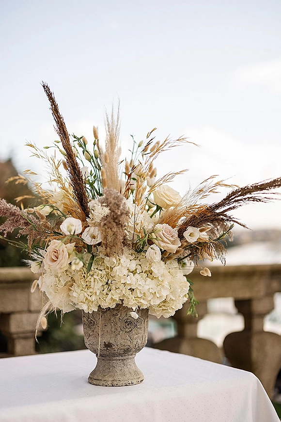 Wedding floral arrangement in a stone urn with white hydrangeas, roses, and pampas grass on a terrace overlooking water and sky