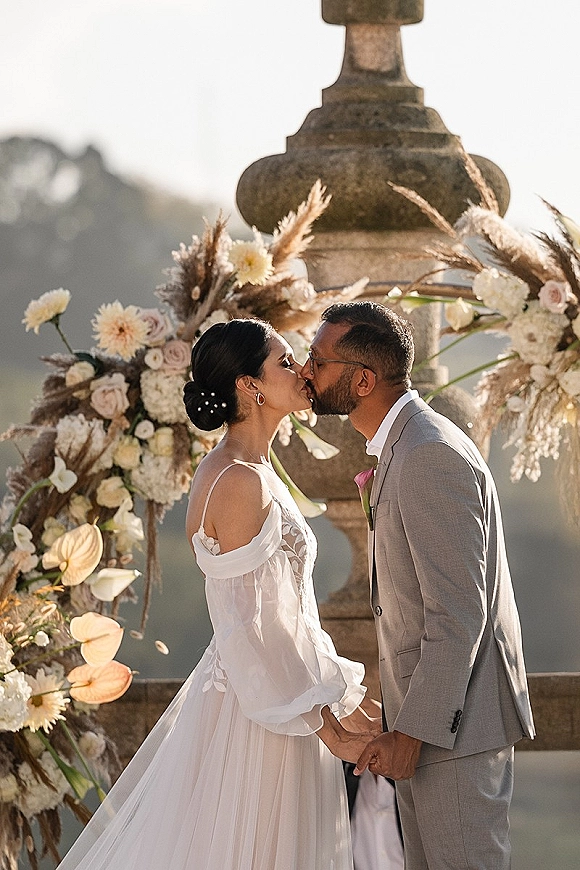 Wedding kiss beneath ceremony kiss moment arch with pampas grass and blush roses on a stone terrace with mountains in the background