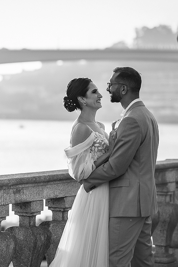 Couple portrait of a wedding couple hugging by a stone balustrade, bride in off-the-shoulder dress, groom in glasses, with water behind