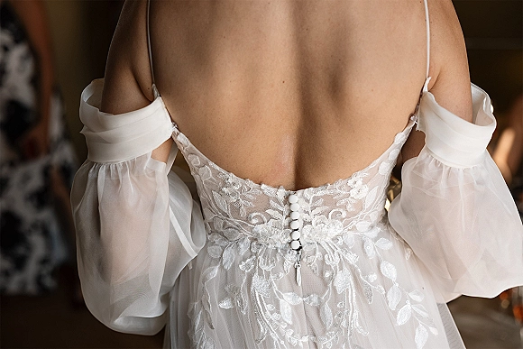 Wedding dress back with illusion lace and fabric-covered buttons, off-the-shoulder sheer tulle sleeves in a softly blurred indoor room