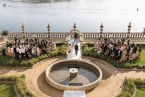 Outdoor wedding ceremony on a waterfront terrace with a white aisle runner leading to a floral ceremony arch and seated guests, overhead view