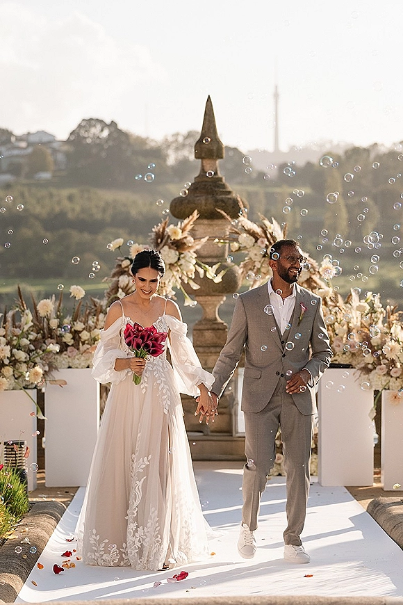 Wedding recessional as bride and groom walk hand in hand through bubbles, bride holding calla lilies on a terrace with stone fountain backdrop