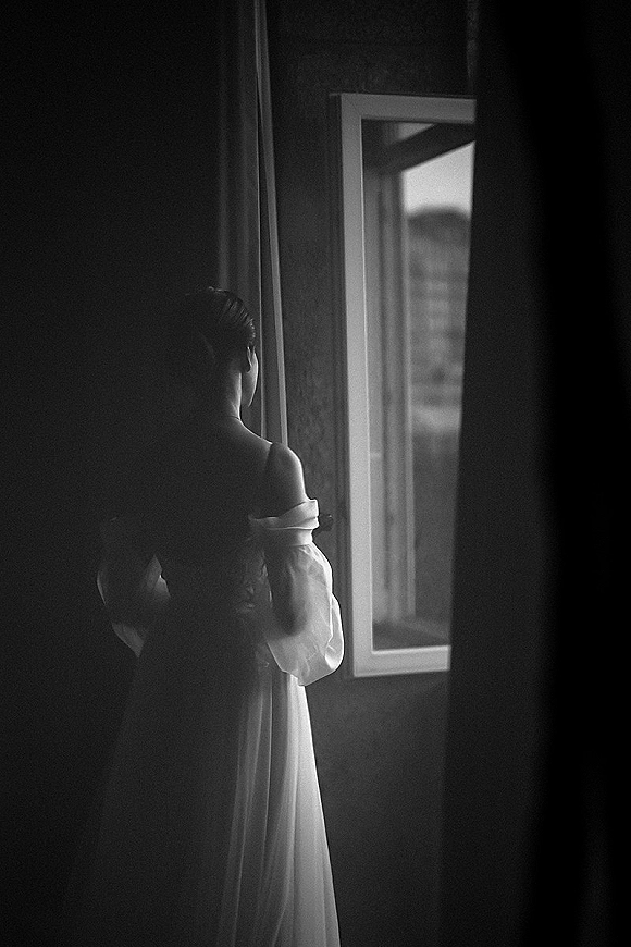 Bridal portrait of a bride by window in soft natural light, showing off-the-shoulder gown with sheer puff sleeves beside curtains