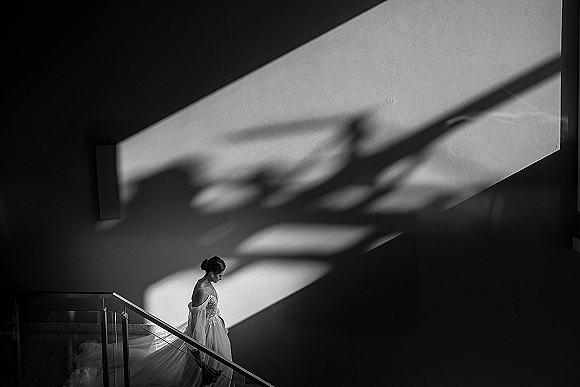 Bridal portrait of a bride on staircase, looking down as her veil and dress train fall in dramatic window light by a glass railing