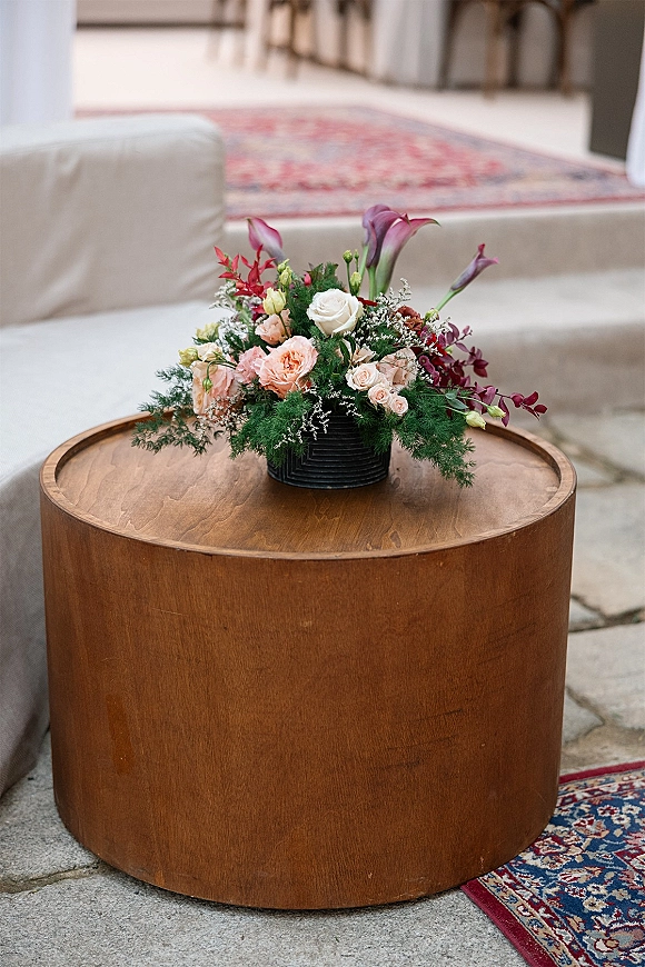 Wedding centerpiece in a black vase with low floral centerpiece roses, calla lilies, and greenery on a round wooden table in a lounge setting