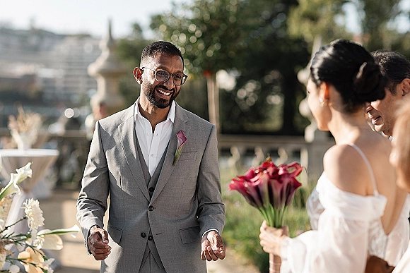 Wedding ceremony moment as groom in gray suit and glasses smiles at bride in white dress holding bouquet by garden fountain and stone balustrade