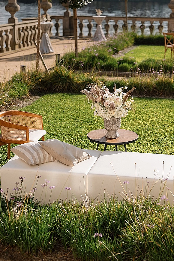 Lounge seating decor with a white sofa, throw pillows, and urn floral centerpiece on a garden lawn by a stone balustrade and water