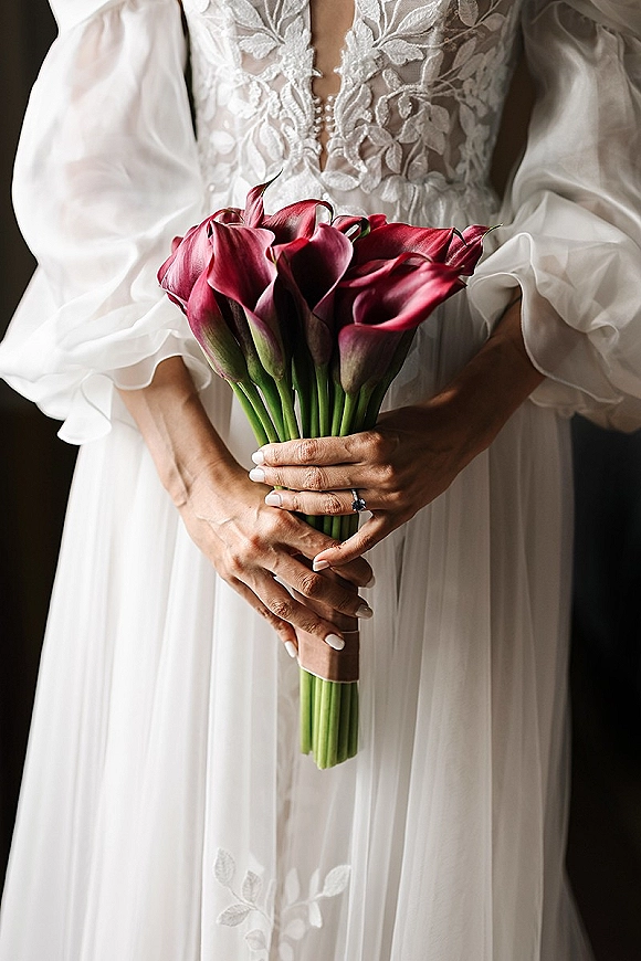 Bridal bouquet of calla lilies held in manicured hands, engagement ring visible against a white lace dress in a dark indoor setting