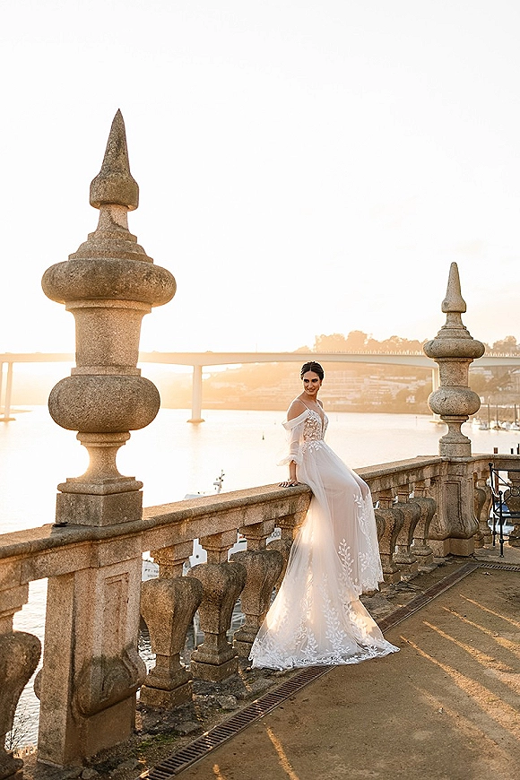 Bridal portrait of a bride sitting on a stone ledge in a lace bodice gown, overlooking a river, bridge, and sunset skyline
