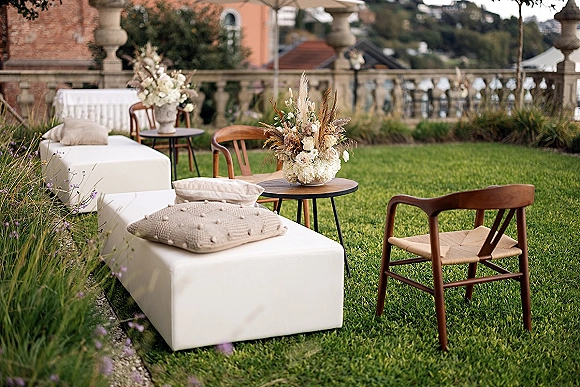Wedding lounge area with white ottomans and pillows around round coffee tables, pampas grass centerpieces on a stone terrace lawn