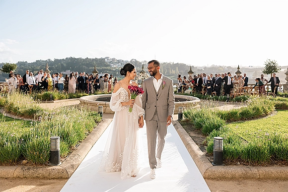 Recessional moment as bride and groom walk the aisle holding hands, bride with burgundy calla lily bouquet on white runner in garden lawn