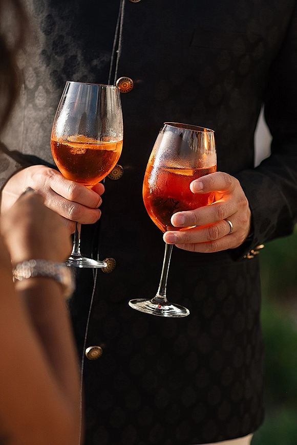 Wedding toast as couple clinking glasses, showing orange cocktail and wine glasses with wedding bands against soft outdoor greenery blur