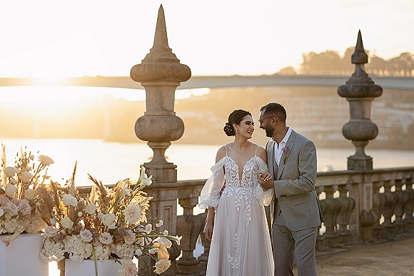 Couple portrait at sunset, bride in lace dress holding groom’s arm on a stone terrace with river and bridge behind, pampas grass accent