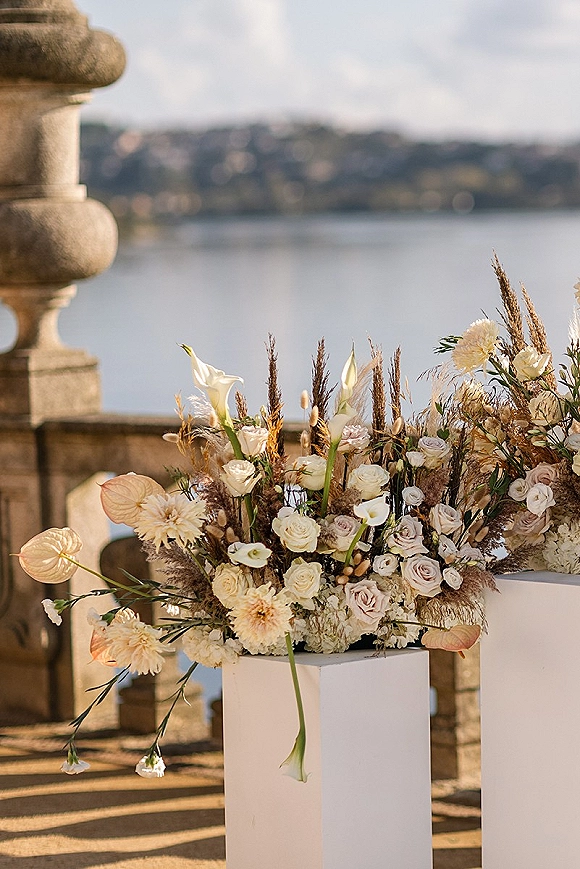 Wedding floral arrangement of calla lilies, roses, dahlias and anthurium on a white pedestal, set by a lakeside stone balustrade
