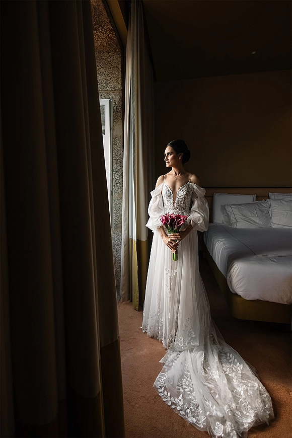 Bridal portrait of a bride holding bouquet of pink tulips in an off-the-shoulder lace gown by a hotel room window with curtains