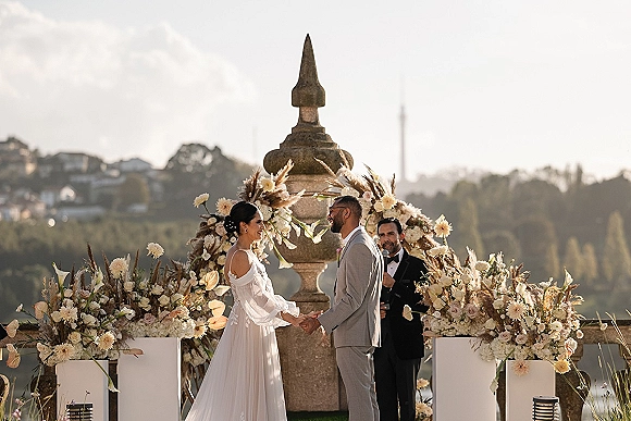 Wedding vows as bride and groom hold hands before a circular floral arch on a stone terrace with hills and water view beyond