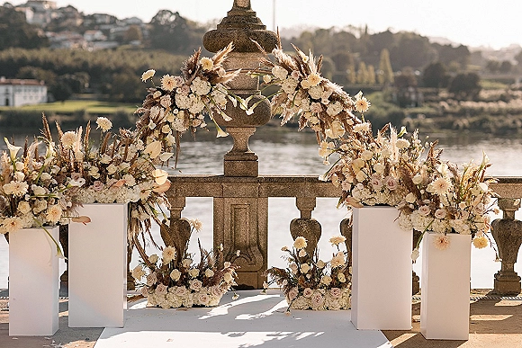 Ceremony backdrop featuring a wedding floral arch with pampas grass on white pedestal plinths beside a stone balustrade overlooking the waterfront