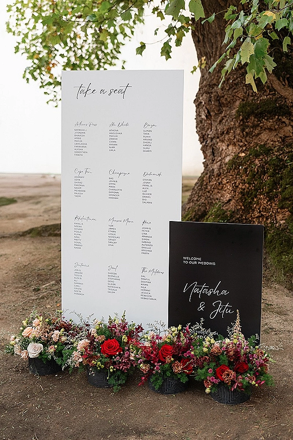 Wedding seating chart with take a seat sign in calligraphy, framed by floral arrangements in black ribbed planters on a lakeside path