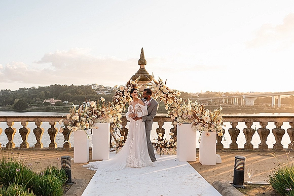 Couple portrait of bride and groom embracing in a lace-sleeve wedding dress and gray suit under a floral arch by the waterfront at sunset