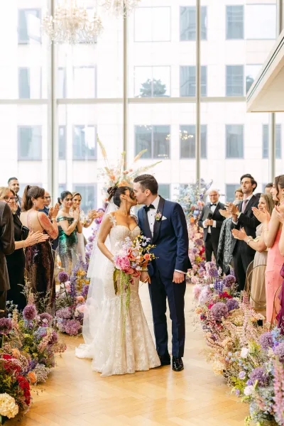 Wedding kiss as bride and groom kiss in a floral-lined aisle, bouquet and veil beneath a chandelier with large windows and guests applauding