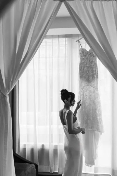 Getting ready bride in a silk slip admiring a lace wedding dress on a hanger by a large hotel room window with sheer curtains