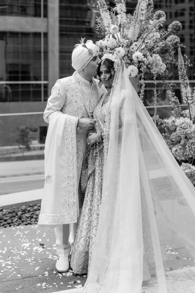Couple portrait of a South Asian wedding couple sharing a forehead kiss, bride in embroidered lehenga and veil by a modern facade water feature