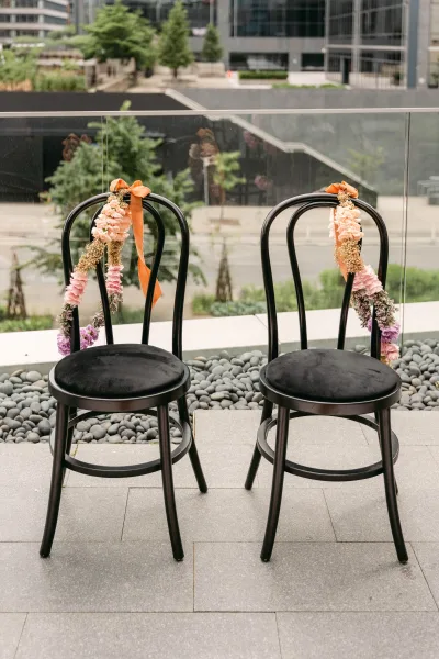 Ceremony chair decor with wedding chair florals, pink and dried garlands tied with orange ribbon on black chairs by a glass railing terrace