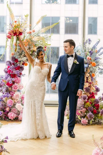 Couple portrait of bride holding bouquet up with groom holding hands, framed by a wedding floral backdrop near large windows and city view