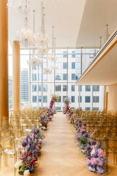 Ceremony aisle design with flower lined wedding aisle, floral pillars and clear acrylic chairs under crystal chandeliers by city-view windows