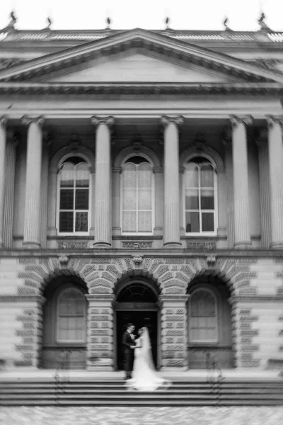 Couple portrait of bride and groom holding hands, her veil train flowing, posed on stone steps before a grand columned doorway