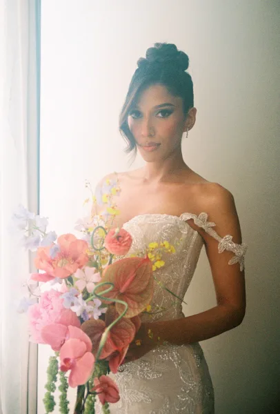 Bridal portrait of a bride in a strapless lace wedding dress holding anthurium and pink pastel bouquet by soft window light on a neutral wall