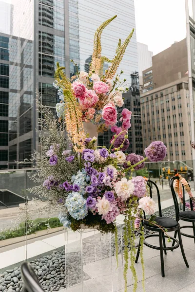 Ceremony floral arrangement of pink peonies, blue hydrangea, and orchids on clear acrylic pedestal on a rooftop terrace with city skyline