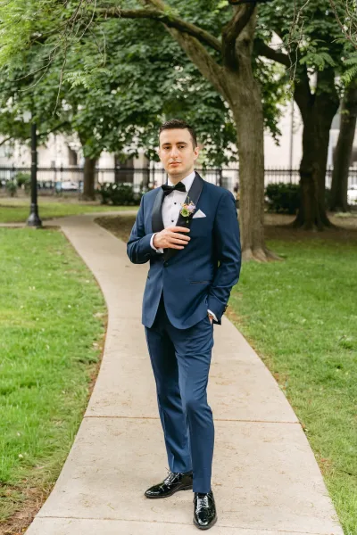 Groom portrait in a navy tuxedo with black bow tie and boutonniere, standing on a park path with trees, fence, and lamppost behind.