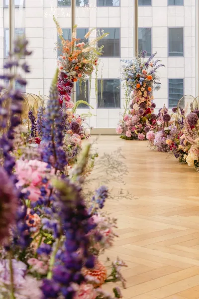 Ceremony altar decor with a floral ceremony arch of purple and pink blooms, hanging greenery, and clear ghost chairs by tall windows