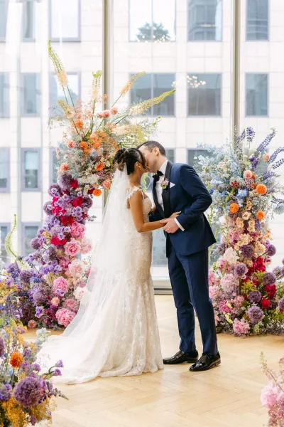Wedding kiss portrait of bride and groom kissing beneath a floral arch, her veil and lace gown framed by large city windows indoors
