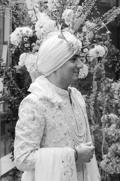 Groom portrait of a Sikh groom in an embroidered ivory sherwani and jeweled turban brooch, posed before a floral greenery backdrop near a window frame
