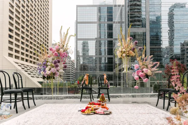 Wedding ceremony setup with pastel florals and black chairs on a rooftop terrace, hanging blooms framing a glass railing and city skyline