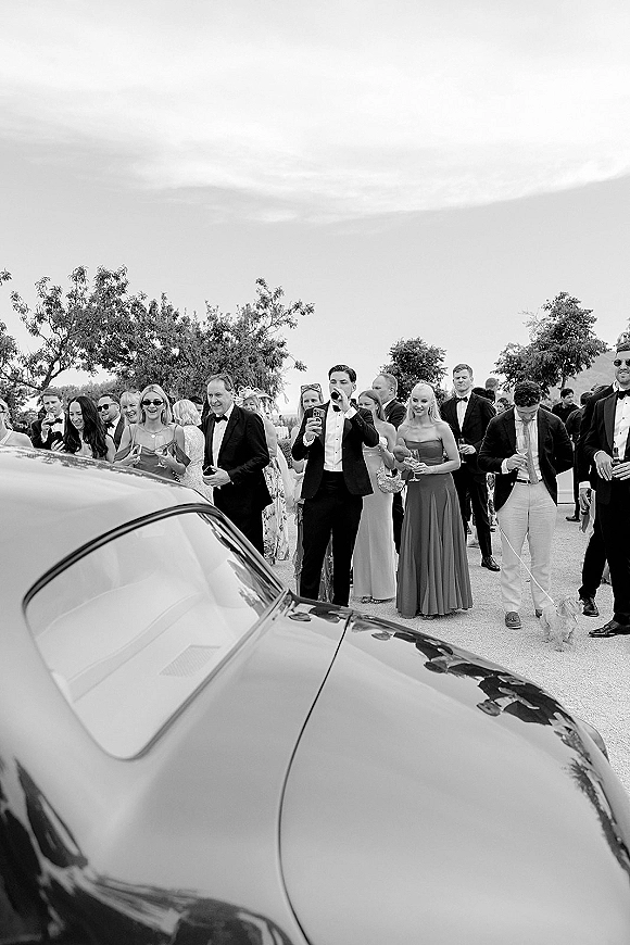 Wedding guest candid of wedding guests cheering in tuxedos and satin dresses, holding champagne flutes by a classic car on a tree-lined driveway