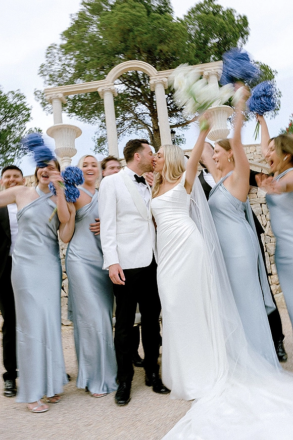 Wedding party portrait of the bride and groom kissing as bridesmaids hold bouquets and cheer on stone steps beneath a columned arch