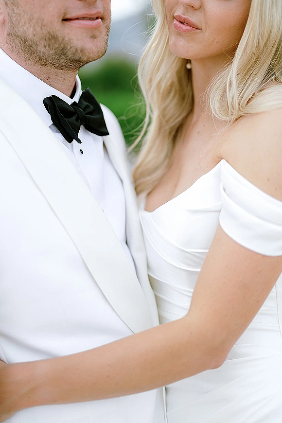 Couple portrait of bride hugging groom in a close up couple portrait, off-shoulder wedding dress against blurred outdoor greenery backdrop