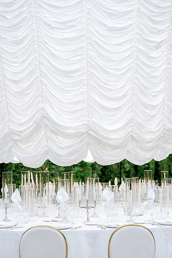 Reception tablescape with white wedding tablescape linens, taper candles and candelabras beneath a draped canopy in a garden setting