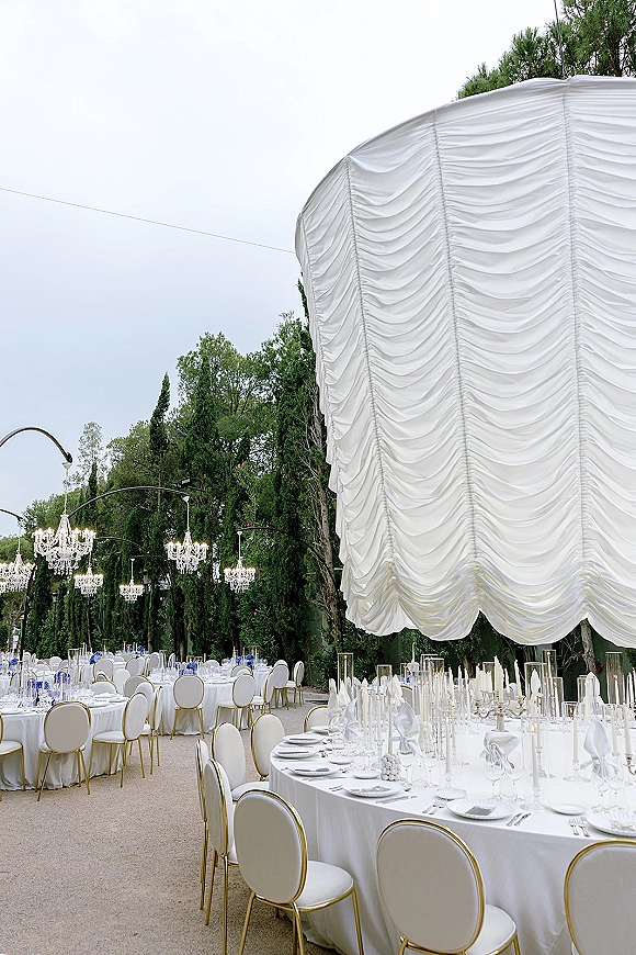 Outdoor reception decor under a draped tent ceiling with crystal chandeliers, round tables, taper candles, and blue florals in a garden setting