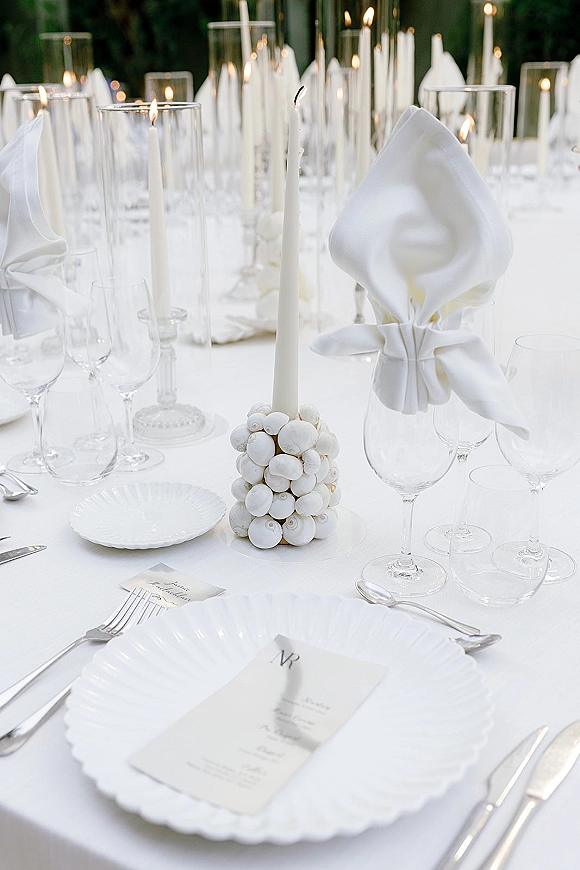 Reception tablescape with an all white tablescape of scalloped plates, folded napkins, menus, place cards, and taper candles amid greenery