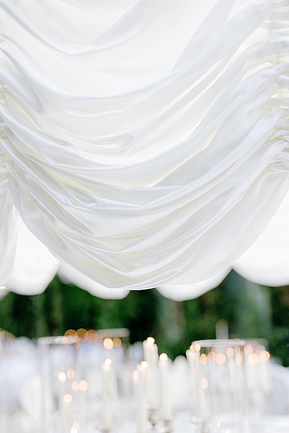 Reception ceiling draping in a white fabric canopy above candlelit tables with taper candles in glass holders, framed by trees outdoors