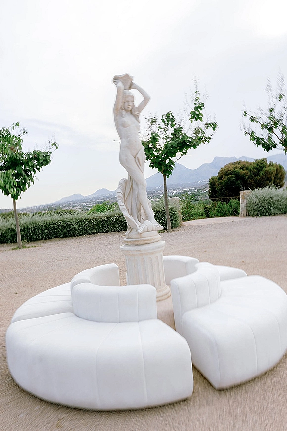 Lounge seating in a wedding lounge area with white modular sofas around a statue pedestal on a gravel patio, mountains beyond hedges