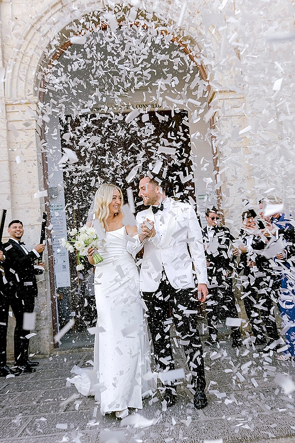 Wedding recessional with newlyweds walking out hand in hand as guests toss confetti under a stone arch, bride in veil and bouquet, groom in white jacket