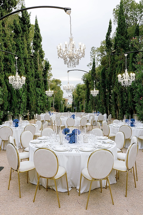 Outdoor reception setup with round wedding tables in white linens, blue floral centerpieces, and crystal chandeliers along a tree-lined path